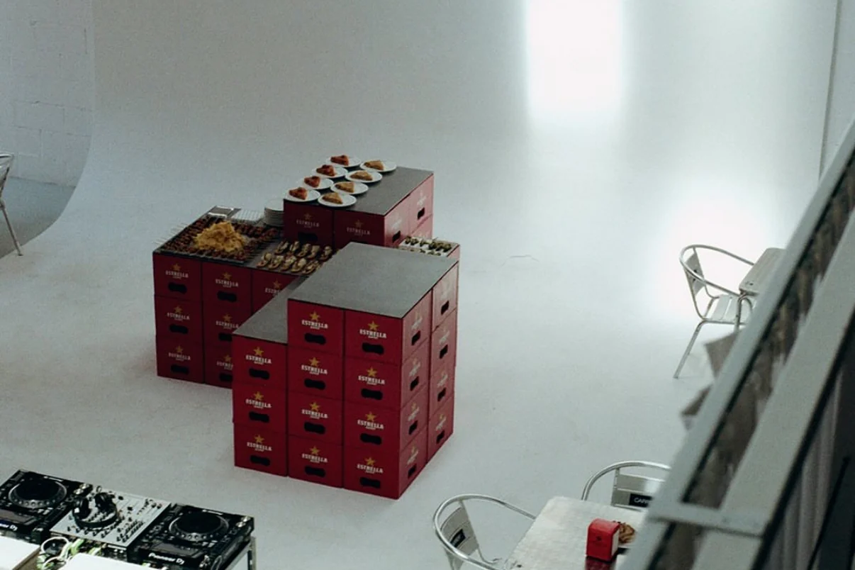 La sala desde arriba: cajas de Estrella Damm apiladas como mesa de catering, sillas metálicas y, en la esquina, la cabina con los CDJ